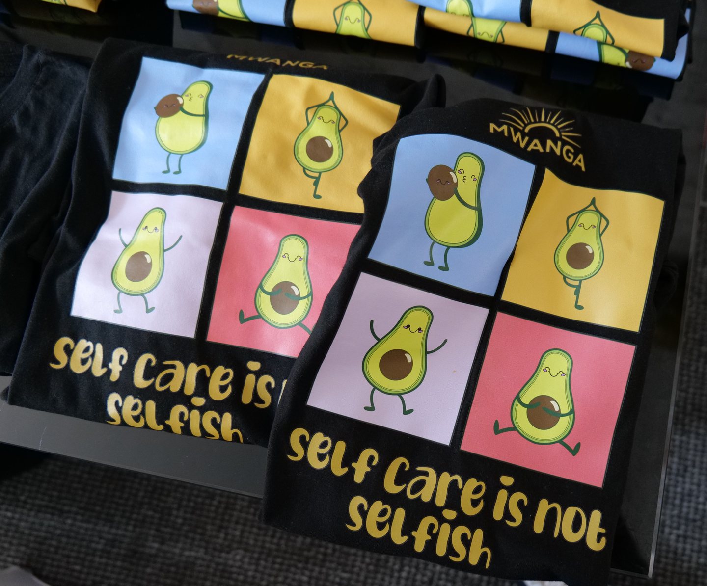 Mwanga T-shirts printed with cartoon avocados and the words self care is not selfish, beneath the Mwanga sunrise logo.